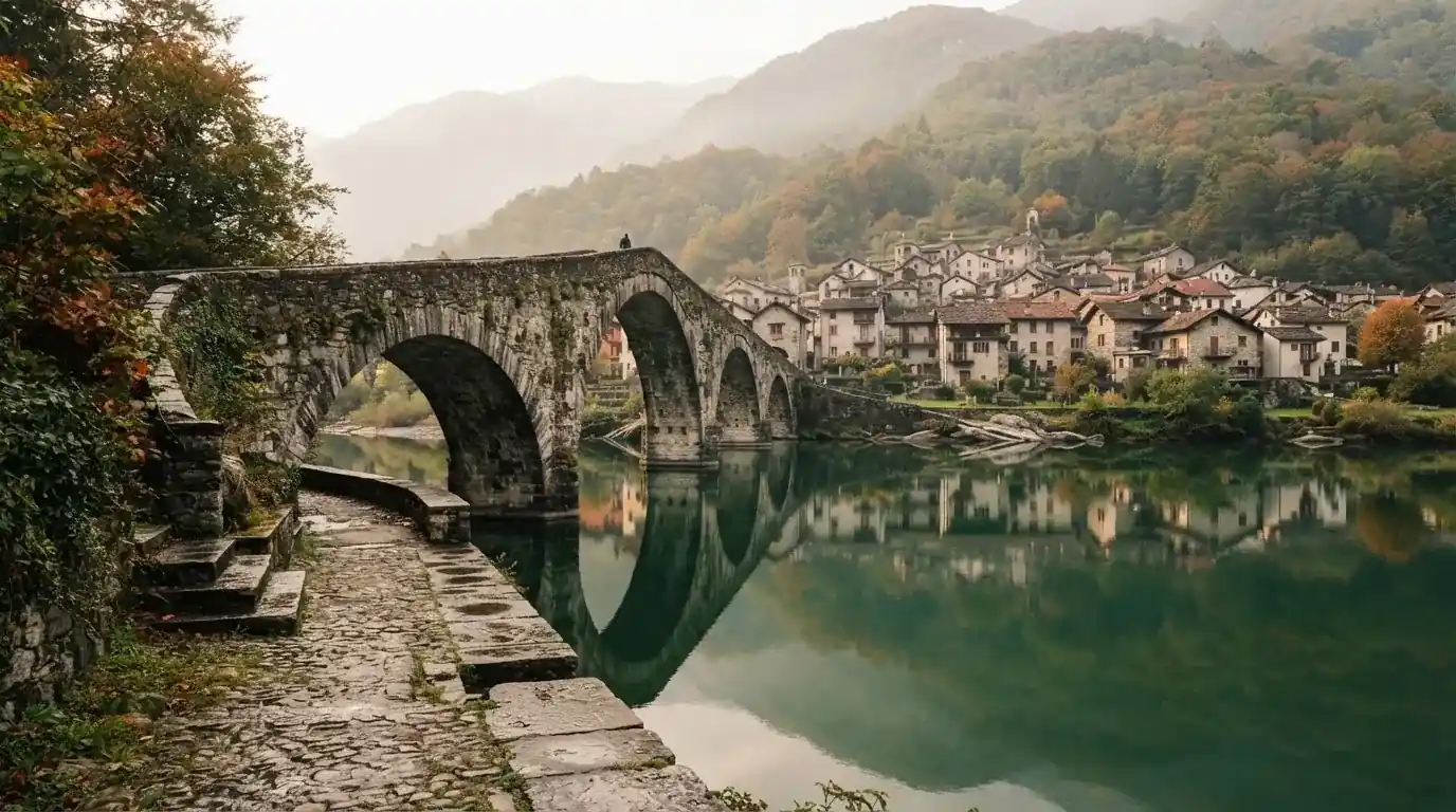 Eine alte Steinbrücke über einen ruhigen Fluss, dahinter Häuser mit Ziegeldächern und bewaldete Hügel.