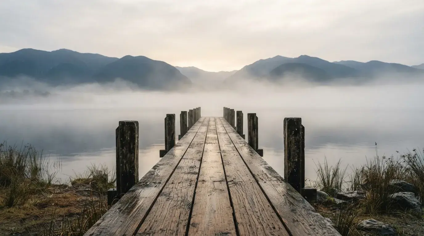 Ein Seeufer mit einem hölzernen Steg, Nebel liegt über dem Wasser, am anderen Ufer erheben sich Berge.