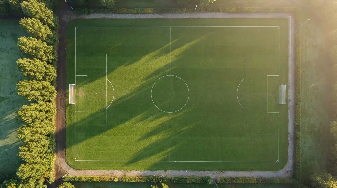 Ein leerer Fussballplatz mit weissen Linien von oben gesehen im Morgenlicht, am Rand stehen zwei Tore und eine Reihe Pappeln.