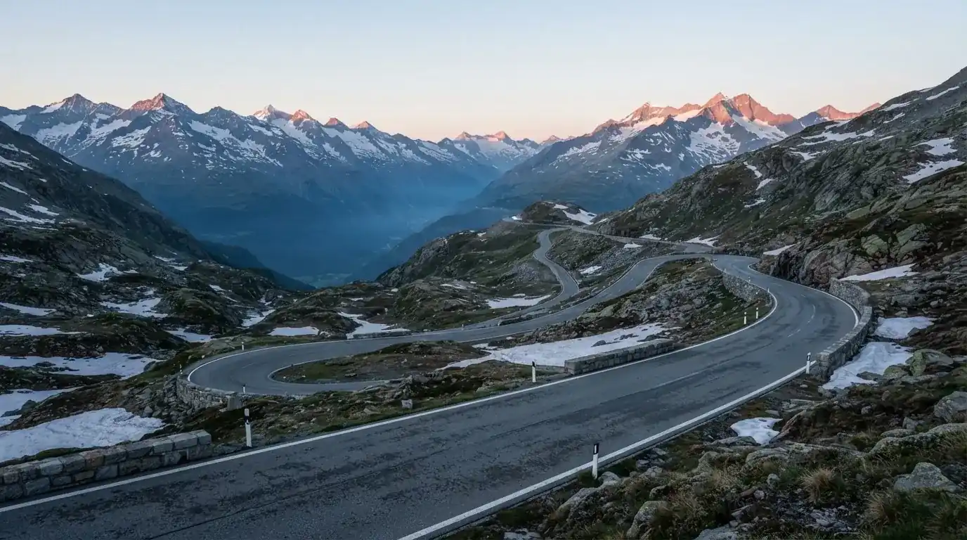 Ein Bergpass in den Schweizer Alpen mit leerer Strasse und Schneeresten am Rand im ersten Frühlicht.