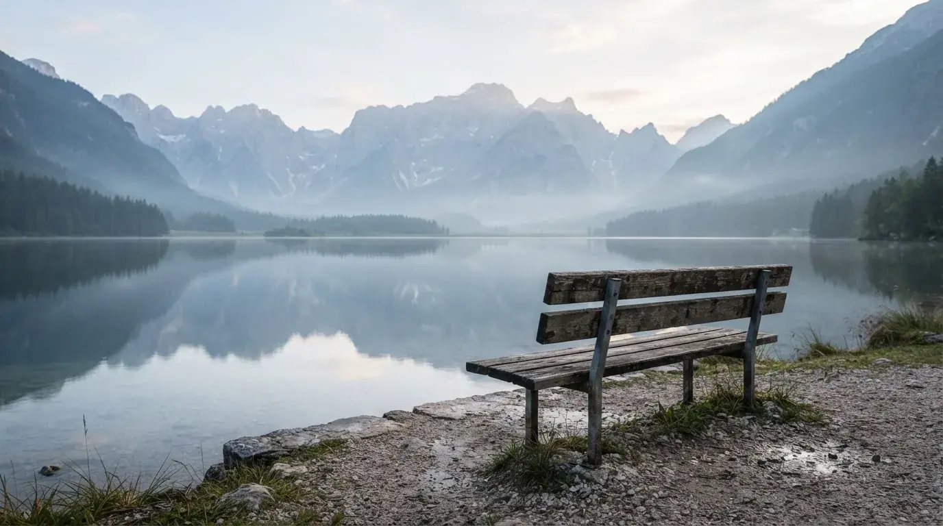 Eine Holzbank an einem Seeufer mit Blick auf eine Bergkette im Morgendunst, das Wasser ist spiegelglatt.