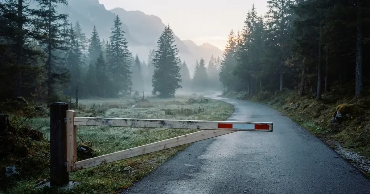 Eine geschlossene Schranke steht an einer ruhigen Bergstrasse im Morgennebel, dahinter Tannen und eine nasse Wiese.