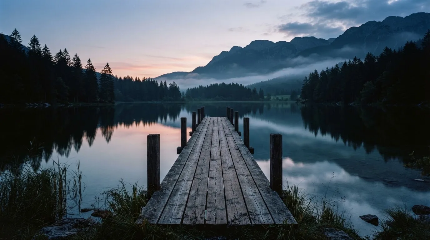Ein Bergsee in der Dämmerung mit einem leeren Holzsteg, die Wasseroberfläche ist ruhig und Nebel zieht über die Hänge.