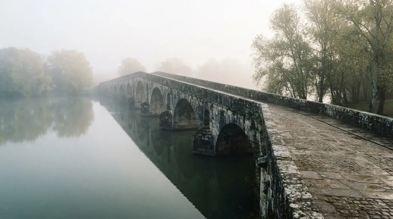 Eine Steinbrücke führt im Frühnebel über einen Fluss, auf dem Fussweg ist niemand zu sehen.